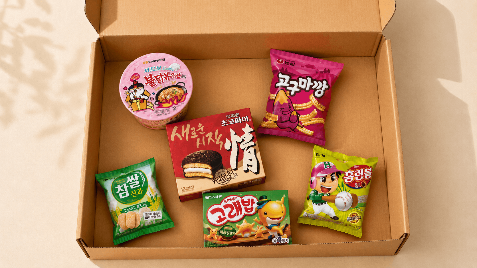 A brown kraft shipping box on a cream tabletop, arranged with iconic Korean snacks — Buldak ramen, Choco Pie, sweet potato puffs, songpyeon, and seaweed-rice snacks.